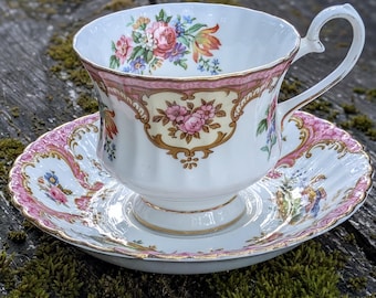 May include: A white porcelain teacup and saucer set featuring a delicate floral pattern with pink, yellow, and green accents. The rim of both pieces is decorated with a scalloped pink border and gold trim, with additional floral motifs inside the cup.