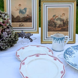 K&ouml;nnte beinhalten: Zwei gerahmte antike Drucke von Frauen in Vintage-Kleidung, einer mit einer Frau in einem blauen Kleid und der andere mit zwei Frauen in einer Gartenlandschaft. Die Drucke werden auf einer wei&szlig;en Tischdecke mit zwei wei&szlig;en Tellern mit rotem Rand, einem blau-wei&szlig;en Krug und einem blau-wei&szlig;en floralen Teller ausgestellt.
