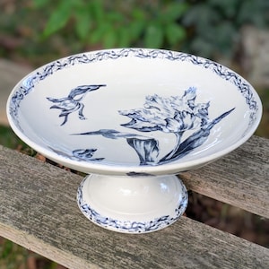 Peut inclure: Plat à pied en céramique blanche avec un motif floral et d'oiseaux bleu. Le plat a un bol large et peu profond et un bord décoratif. La base est ronde et également décorée d'accents bleus. Le motif représente une grande fleur et un oiseau.
