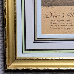 K&ouml;nnte beinhalten: Ein goldfarbener Bilderrahmen mit einem wei&szlig;en Passepartout und einem braunen Papiereinsatz. Der Einsatz enth&auml;lt eine handschriftliche Inschrift in Franz&ouml;sisch, die lautet "D&eacute;di&eacute;e &agrave; Me Blaise &agrave; la partin l'appr&eacute;te Li meme a faire le guet. Du chapiau couvrant la t&ecirc;te, 1 place au lieu du piquer."