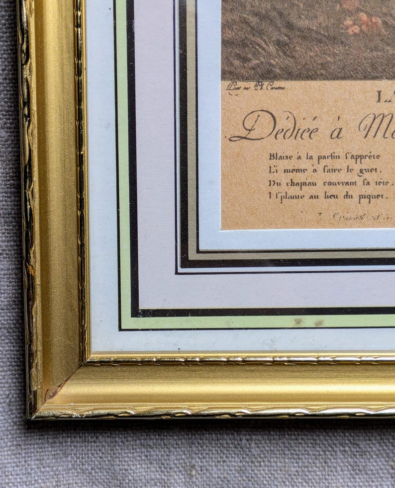 K&ouml;nnte beinhalten: Ein goldfarbener Bilderrahmen mit einem wei&szlig;en Passepartout und einem braunen Papiereinsatz. Der Einsatz enth&auml;lt eine handschriftliche Inschrift in Franz&ouml;sisch, die lautet "D&eacute;di&eacute;e &agrave; Me Blaise &agrave; la partin l'appr&eacute;te Li meme a faire le guet. Du chapiau couvrant la t&ecirc;te, 1 place au lieu du piquer."