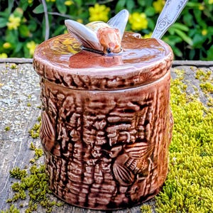 Puede incluir: Un tarro de miel de cerámica marrón con tapa de abeja y cuchara plateada. El tarro tiene un diseño de corteza de árbol texturizado. El fondo presenta flores amarillas y follaje verde.