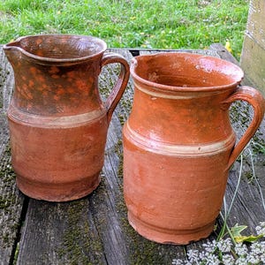 Könnte beinhalten: Zwei rustikale Krüge in Terrakotta-Farbe mit Griffen. Die Krüge haben einen breiten Boden und eine schmalere Oberseite mit Ausguss. Ein helles Band umschließt die Mitte jedes Kruges. Die Krüge werden auf einer verwitterten Holzoberfläche präsentiert.