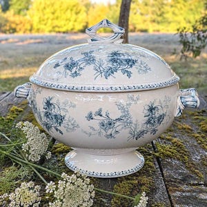 Peut inclure: Une soupière en céramique couleur crème avec son couvercle assorti, ornée d'un motif floral bleu en transfert. La soupière présente deux anses latérales, une base sur pied et un bouton de couvercle décoratif, tous décorés de motifs floraux bleus.