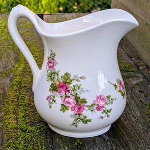 Peut inclure: Pichet en céramique blanche avec une anse incurvée et un motif floral de roses roses et de feuilles vertes. Le pichet a une large ouverture et un corps arrondi, pour servir des boissons.