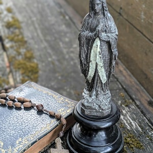 Könnte beinhalten: Eine dunkelgraue Statue einer Figur mit Krone und Gewand, die Hände zum Gebet gefaltet, auf einem schwarzen runden Sockel. Der Sockel der Statue trägt die Aufschrift "N.D. LOURDES". Ein schwarzes Buch mit Goldverzierung und ein hölzerner Rosenkranz.