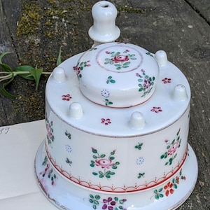 Pode incluir: Recipiente de cerâmica branca com tampa, adornado com rosas cor-de-rosa e folhas verdes. O recipiente tem uma base redonda e um corpo cilíndrico com pequenas protuberâncias brancas e arredondadas. A tampa apresenta um design floral a condizer. Uma peça decorativa.