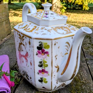 Può includere: Teiera in porcellana bianca con corpo e coperchio quadrati, ornata da filigrana dorata e motivi floreali. La teiera presenta un beccuccio e un manico curvi, con fiori rosa e viola dipinti sul lato. Un panno e un fiore viola sono in primo piano.