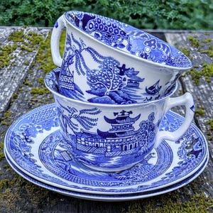 May include: Two white ceramic teacups and a matching saucer featuring a blue willow pattern. The design depicts traditional pagodas, willow trees, and bridges. The teacups have curved handles and the saucer has a wide, decorative blue border.