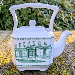 Peut inclure: Théière en céramique blanche avec un motif vert et blanc représentant une devanture de magasin avec le texte "Jacksons Tea Coffee Herbs Spices" sur le devant. La théière a une poignée et un couvercle.