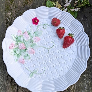 Peut inclure: Une assiette de service en céramique blanche avec un motif de panier tressé et des bords festonnés. L'assiette présente un motif de fraises peintes et est ornée de fraises fraîches et de fleurs.