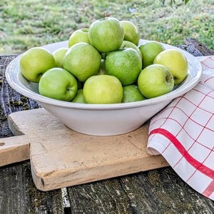 Puede incluir: Un cuenco grande de cerámica blanca lleno de un montón de manzanas verdes frescas. El cuenco se asienta sobre una tabla de cortar de madera junto a un paño blanco con un patrón de cuadrícula roja. Las manzanas varían de verde amarillento a verde.