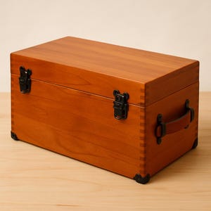 May include: A rectangular wooden storage box with a warm, reddish-brown finish. It features black metal hardware, including hinges, corner protectors, and a clasp. A brown leather handle is attached to the side. The box is likely used for storage.