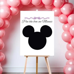Op de afbeelding: Een wit bord op een houten ezel met een zwart silhouet van Mickey Mouses hoofd. Op het bord staat "Pin the bow on Minnie" in script met een paarse strik en lintontwerp. Roze ballonnen vormen een boog en omlijsten het bord.