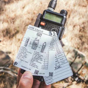 May include: Black and gray handheld radio with a screen and buttons, held with a layout card. The card has diagrams and text, including "UV5R Layout" and "READYRADIO.COM". The background is blurred.