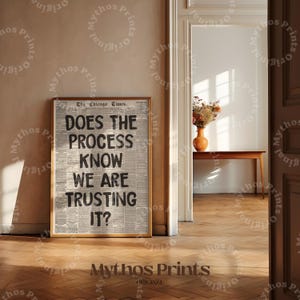 Pode incluir: Uma impressão emoldurada com o texto "DOES THE PROCESS KNOW WE ARE TRUSTING IT?" em letras pretas em negrito. O fundo é uma impressão de jornal. A moldura é de cor castanho claro. A impressão é exibida em uma parede castanho claro.
