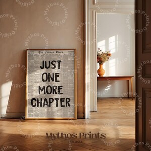 May include: A framed print with the text "JUST ONE MORE CHAPTER" printed in black on a newspaper background. The print is hanging on a wall in a room with wooden floors and a doorway.