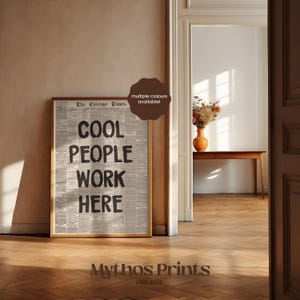 May include: A rectangular framed wall art print featuring a vintage newspaper background with the bold black text "COOL PEOPLE WORK HERE" centered on the page. The print is displayed in a thin wooden frame and includes a brown circular badge.