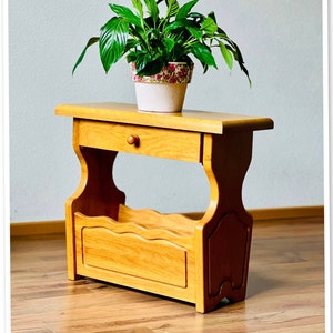 May include: A light wood shelf unit with a drawer and a lower shelf. The shelf is decorated with a wavy design. A potted plant with green leaves sits on top of the shelf.