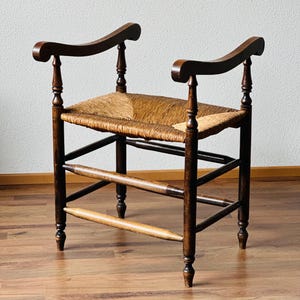 May include: A dark brown wooden chair featuring a woven rush seat and curved armrests. The chair has four turned legs connected by horizontal stretchers, with a low back design and a natural fiber seat texture that contrasts with the dark wood frame.