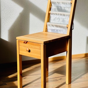 Op de afbeelding: Een houten stoel met een lade en een opbergvak van stof aan de rugleuning. De stoel is gemaakt van licht hout en heeft een eenvoudig ontwerp.