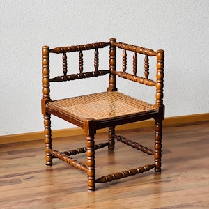 Puede incluir: Una silla de esquina de madera con un asiento de caña tejido y patas torneadas. La silla está hecha de madera marrón oscura y tiene un diseño simple y tradicional.