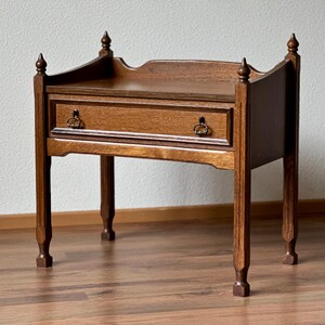 Op de afbeelding: Een bruine houten bijzettafel met één lade voorzien van twee donkere metalen ringgrepen. De tafel heeft vier rechte poten met decoratieve knoppen op de bovenhoeken en een verhoogd achterpaneel met een gebogen rand aan de bovenzijde.