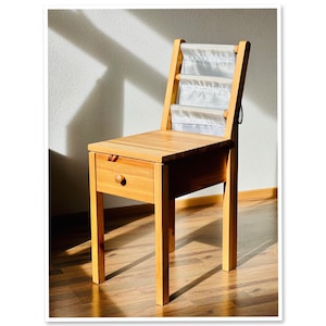 Puede incluir: Una silla de madera con un estante de almacenamiento integrado y un cajón. La silla está hecha de madera clara y tiene un respaldo de tela blanca.