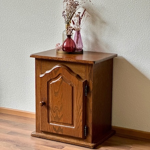 Op de afbeelding: Een kleine houten kast met een enkele deur en een decoratief voorpaneel. De kast is gemaakt van warm getint hout en heeft een kleine, ronde knop. De bovenkant van de kast bevat drie kleine vazen met gedroogde bloemen.