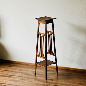 Könnte beinhalten: Ein hoher, hölzerner Pflanzenständer mit einer quadratischen Oberseite und einem Regal in der Mitte. Der Ständer ist aus dunklem Holz gefertigt und hat ein schlichtes, elegantes Design.