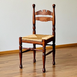 May include: A wooden chair with a medium brown finish, featuring a woven rush seat and a backrest composed of two horizontal slats. The frame includes turned legs and vertical supports topped with rounded finials, connected by thin stretchers.