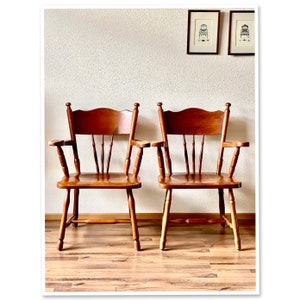 Op de afbeelding: Twee houten stoelen met een lichtbruine afwerking en een eenvoudig ontwerp. De stoelen hebben een gebogen rugleuning, een brede zitting en vier poten. De stoelen staan ​​naast elkaar op een houten vloer.