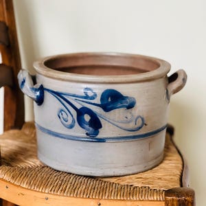 May include: A large, gray stoneware crock with blue floral designs. The crock has two handles and a wide opening.