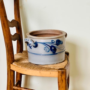 May include: A blue and white glazed ceramic planter with a floral design. The planter has two handles and is sitting on a wooden chair.