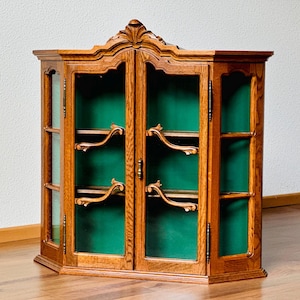 May include: A wooden corner cabinet with glass doors and a green interior. The cabinet has a decorative top and curved shelves. The wood is a warm brown color, and the cabinet is set on a wooden floor.