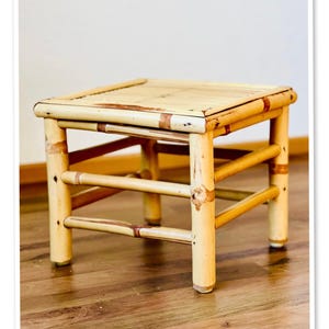 Puede incluir: Un pequeño taburete cuadrado de bambú. El taburete es de color marrón claro con acentos marrones más oscuros. Las patas y el marco están hechos de cañas de bambú, y la parte superior es una superficie plana. El taburete está diseñado con una estética simple y natural.