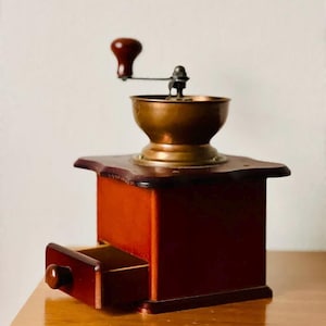 Op de afbeelding: Een vintage houten koffiemolen met een koperen maalcompartiment en een houten lade. De molen heeft een kruk met een rode knop.