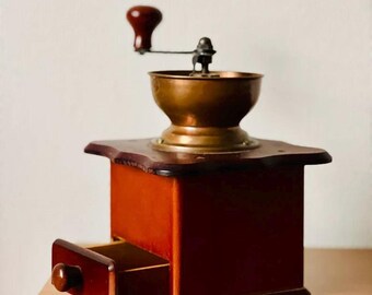Könnte beinhalten: Eine antike Kaffeemühle aus Holz mit einem Mahlwerk aus Kupfer und einer Schublade aus Holz. Die Mühle hat einen Kurbelgriff mit einem roten Knopf.
