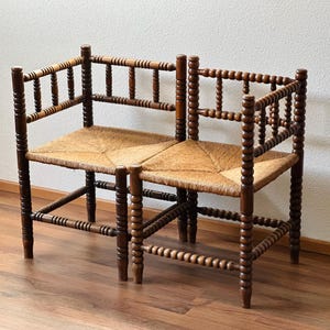 May include: A two-seater wooden bench with a woven straw seat. The bench has a dark brown finish with turned spindles on the legs, back, and arms. The bench is placed on a wooden floor.