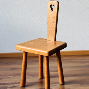 Op de afbeelding: Een kleine houten stoel met een hartvormige uitsparing in de rugleuning. De stoel heeft vier poten en een eenvoudig, rustiek ontwerp.
