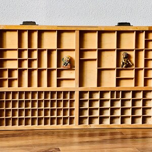 May include: A wooden printing block tray with multiple compartments. The tray is made of light brown wood and has two rows of compartments. The top row has 12 compartments and the bottom row has 24 compartments. There are two small figurines in the top row of compartments.