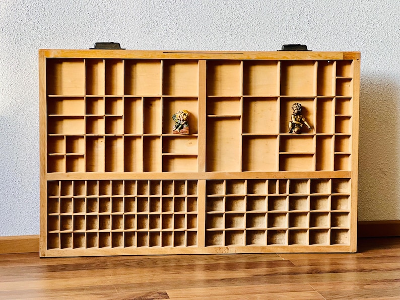 May include: A wooden printing block tray with multiple compartments. The tray is made of light brown wood and has two rows of compartments. The top row has 12 compartments and the bottom row has 24 compartments. There are two small figurines in the top row of compartments.
