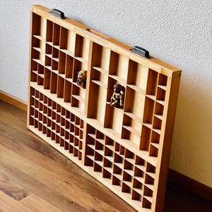 May include: A wooden printer's tray with multiple compartments, featuring a light brown finish and two black metal handles. The tray is empty, but there are two small decorative figurines inside.