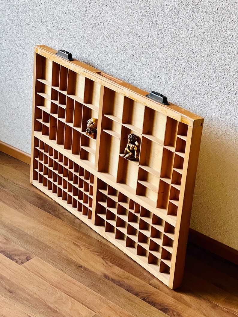 May include: A wooden printer's tray with multiple compartments, featuring a light brown finish and two black metal handles. The tray is empty, but there are two small decorative figurines inside.