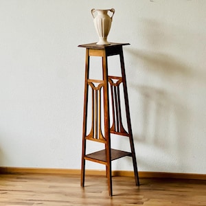 Könnte beinhalten: Ein Pflanzenständer aus Holz mit einer einzigen Ablage und einem verzierten Design. Eine weiße Keramikvase steht auf der oberen Ablage.