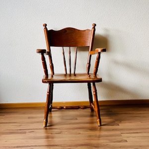 Könnte beinhalten: Ein Holzstuhl mit brauner Oberfläche und einer Lattenrücklehne. Der Stuhl hat eine geschwungene Sitzfläche und Armlehnen. Der Stuhl steht auf einem Holzboden.