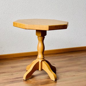 Op de afbeelding: Een kleine, achthoekige houten tafel met een enkele voet en drie poten. De tafel is gemaakt van licht hout en heeft een natuurlijke afwerking.