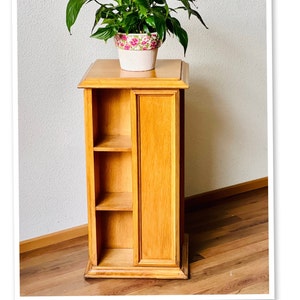 May include: A wooden plant stand with three shelves and a closed cabinet door. The stand is made of light wood and has a simple design. A potted plant with green leaves is sitting on top of the stand.
