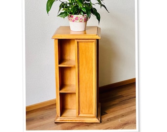 May include: A wooden plant stand with three shelves and a closed cabinet door. The stand is made of light wood and has a simple design. A potted plant with green leaves is sitting on top of the stand.