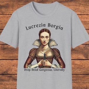 May include: A heather gray t-shirt featuring a graphic of Lucrezia Borgia wearing a high-collared dress and multiple pearl necklaces, holding a small dagger. The text reads Lucrezia Borgia above and Drop Dead Gorgeous. Literally below the figure.